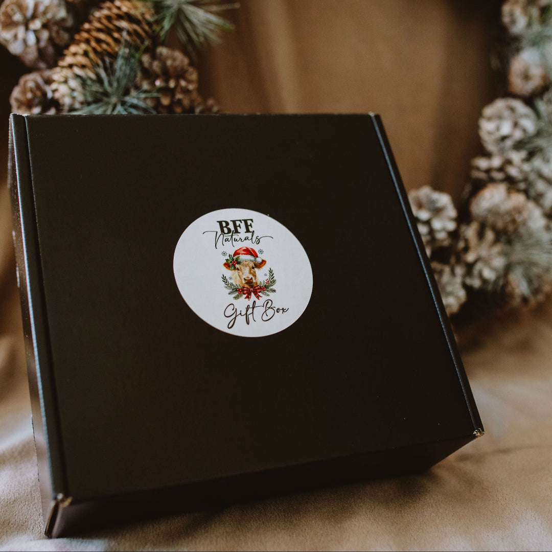Black gift box with a decorative sticker featuring a Santa Claus design, placed against a festive background with pine cones and greenery.