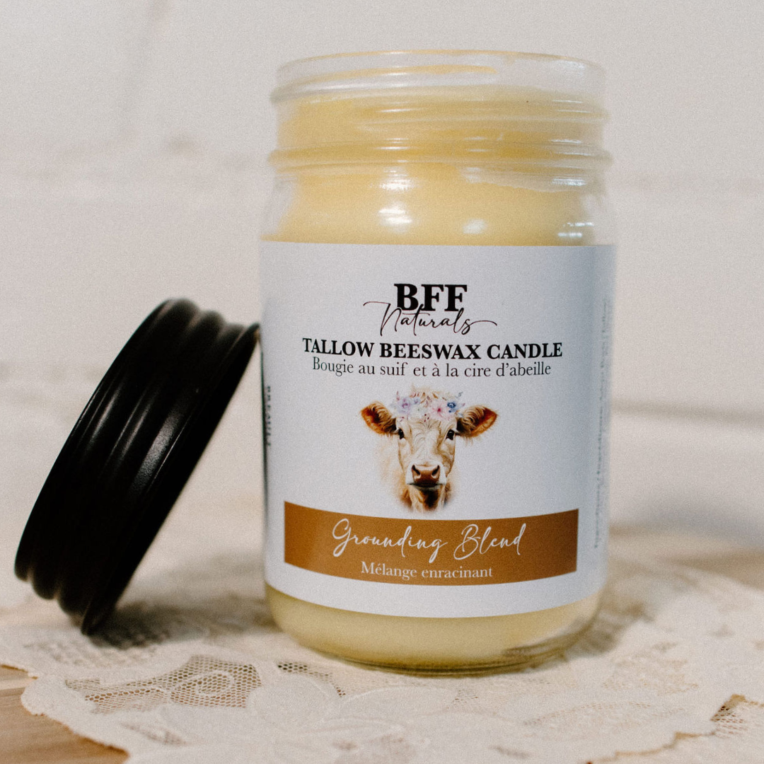 A glass jar candle labeled “BFF Naturals Tallow Beeswax Candle” with the scent name “Grounding Blend / Mélange enracinant.” The label features a cow illustration with a floral crown. The candle lid is black and rests beside the jar on a lace surface.